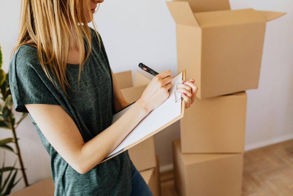 A young woman writes on a clipboard while organizing moving boxes in her home.